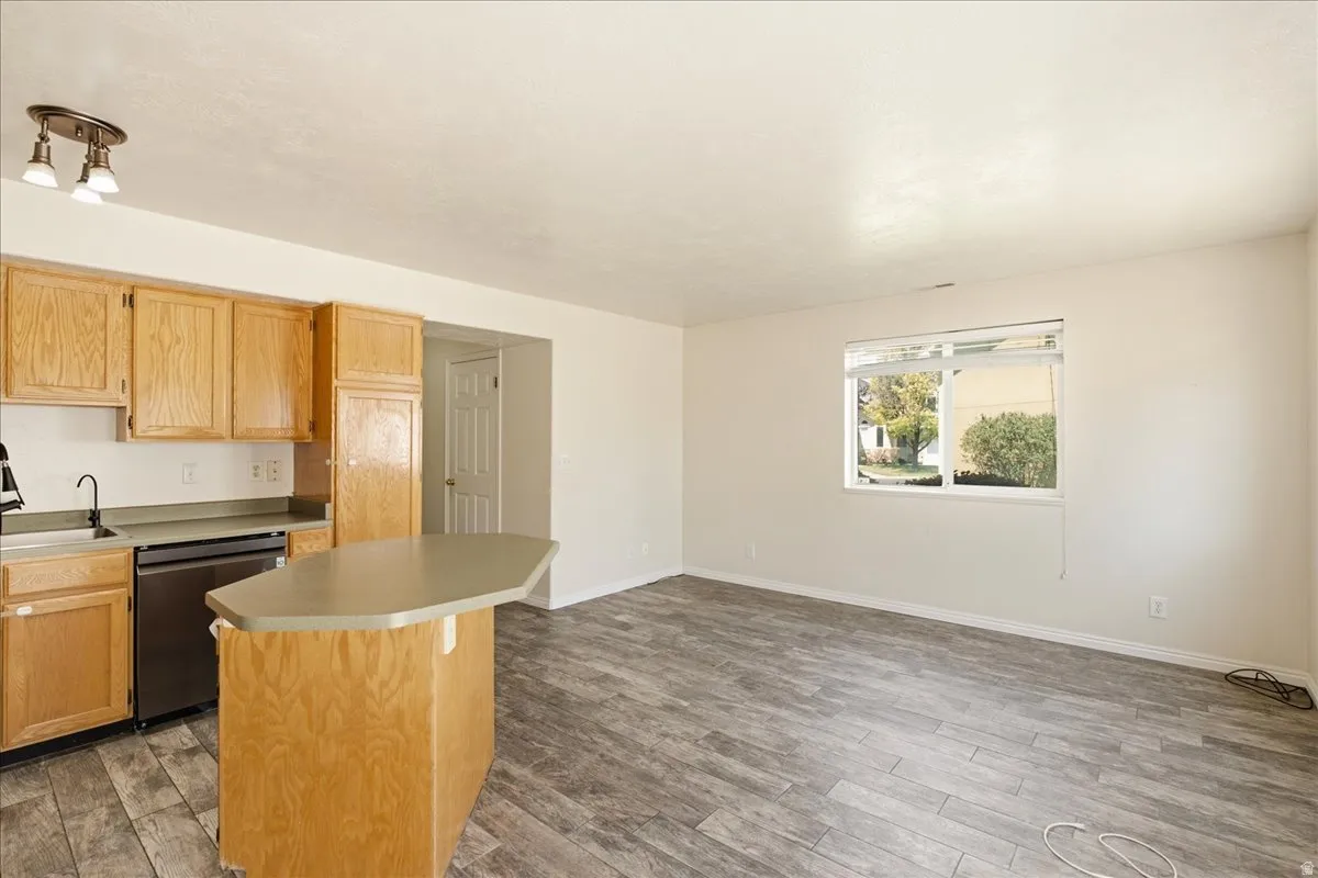Kitchen and living area featuring wood-finish flooring, a kitchen island with seating, natural wood cabinetry, a stainless steel dishwasher, and a window providing exterior views