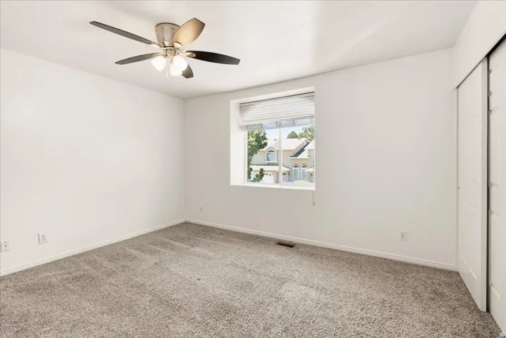 Neutral-toned carpeted interior space featuring a ceiling fan with integrated lighting, a single window with blinds, and white bi-fold closet doors