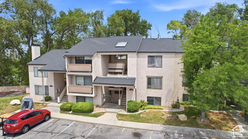 Multi-story residential building with light-colored stucco exterior, dark shingle roofing, multiple balconies, and mature perimeter landscaping
