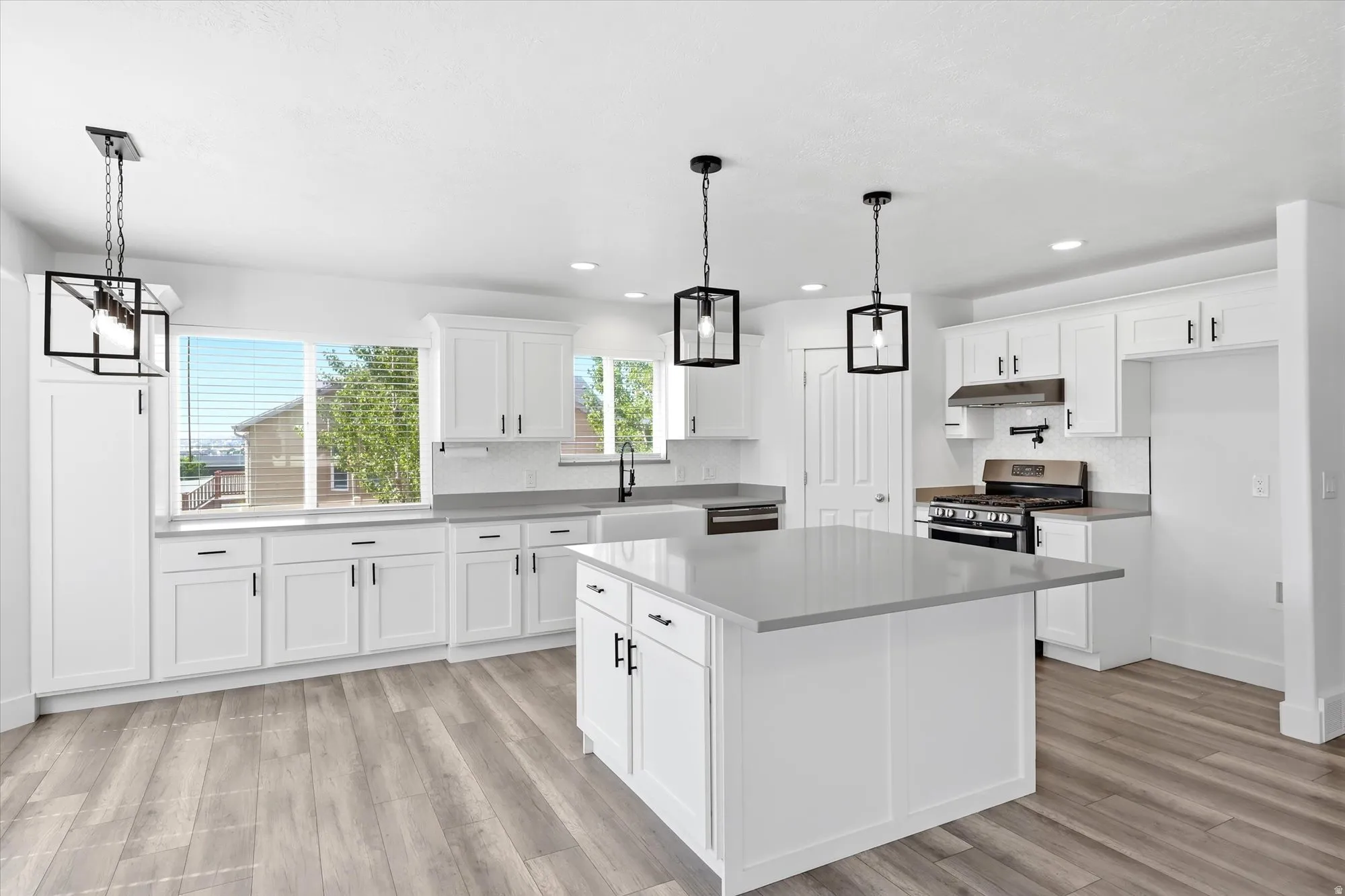 Bright kitchen with white cabinetry, wood-finish flooring, a central island with a light-colored countertop, and modern pendant lighting fixtures