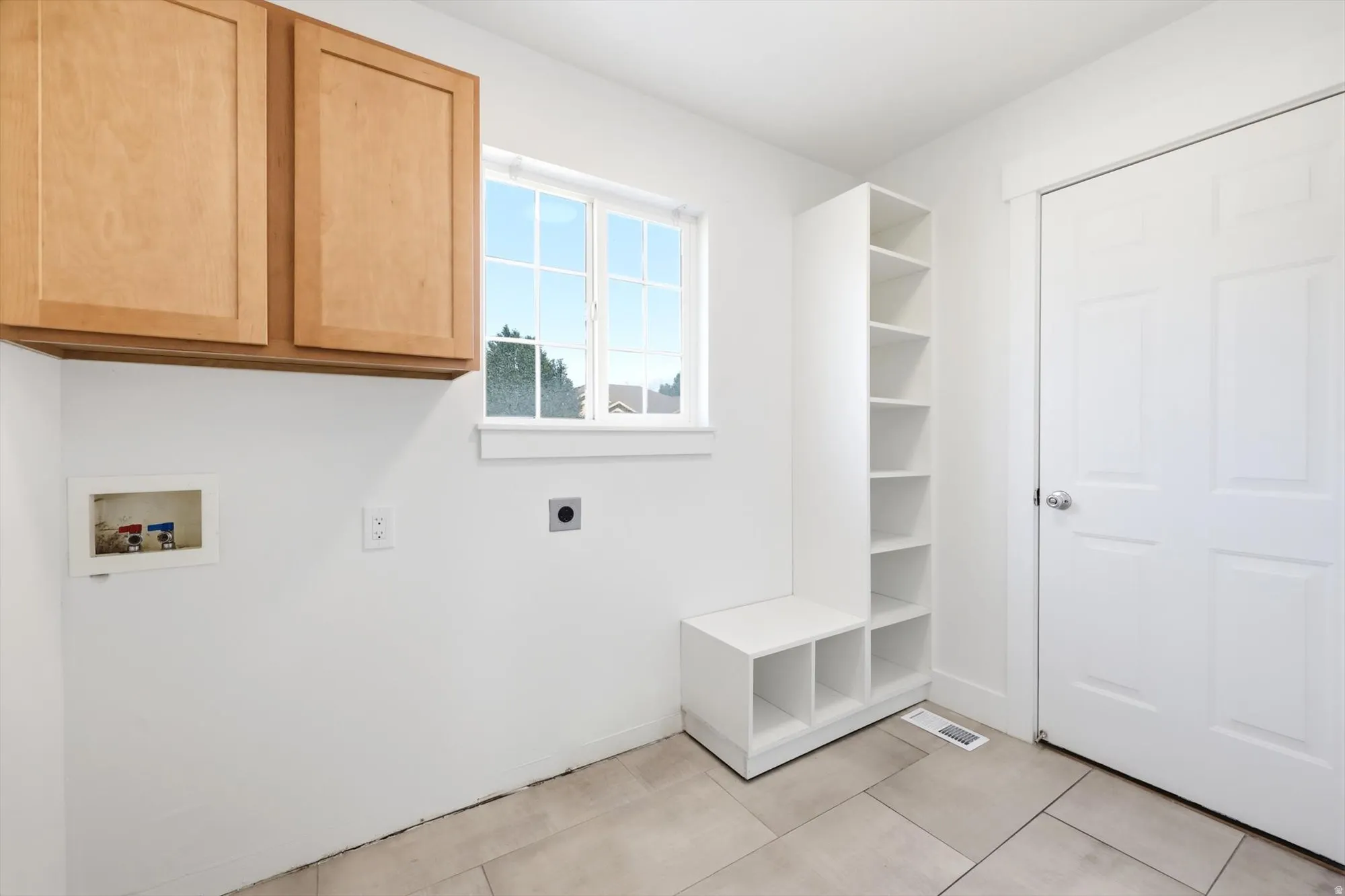 Laundry area featuring light wood-finish cabinetry, a window with grid panes, built-in open shelving, and neutral floor tiling