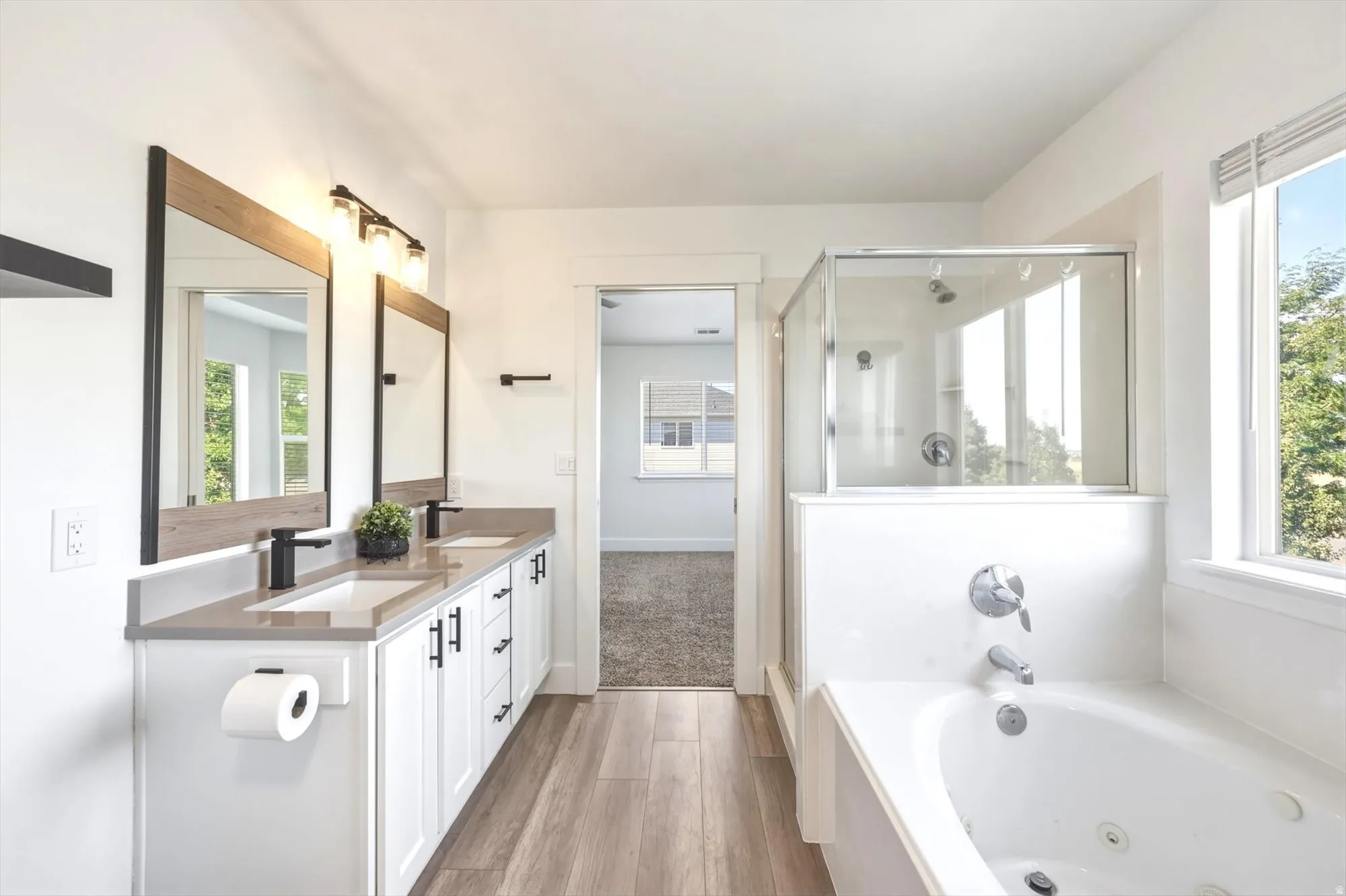 Double vanity featuring rectangular sinks with matte black faucets, wood-finish flooring, a jetted soaking tub, and a separate glass-enclosed shower