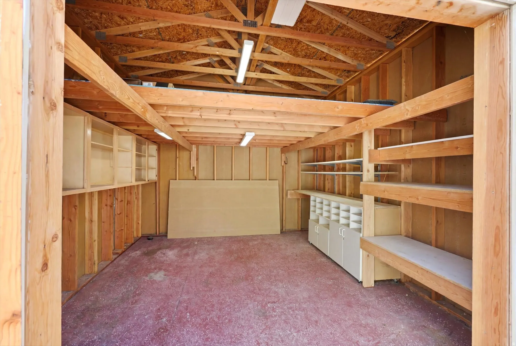 Spacious garage interior featuring exposed wood framing, ample built-in shelving, overhead fluorescent lighting, and a red concrete floor