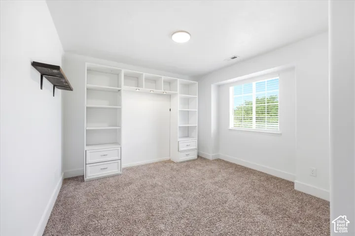 Room featuring plush carpet flooring, a large window with white blinds, and a built-in shelving unit with integrated lighting and drawer storage