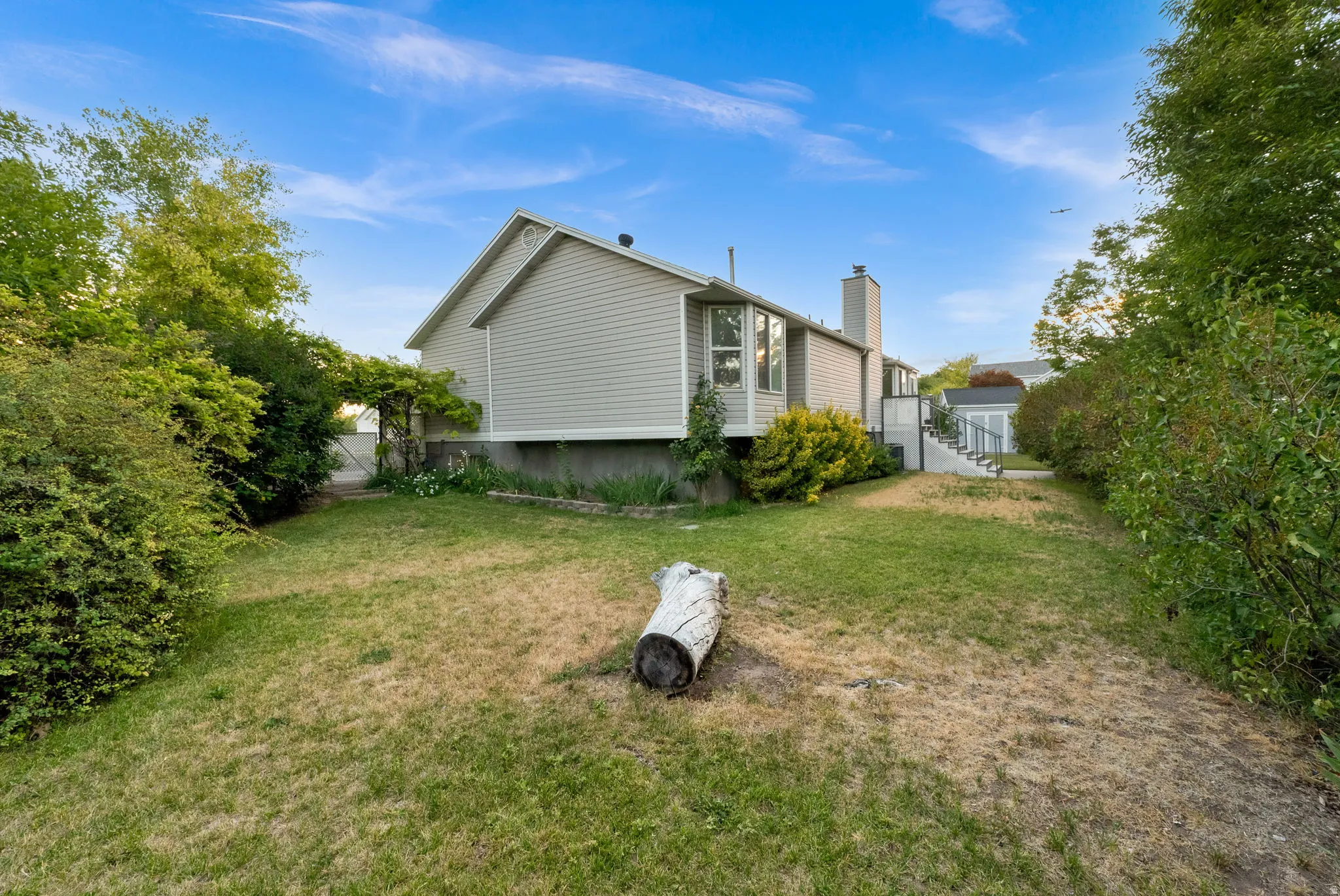 Spacious backyard featuring a sloped lawn area, mature perimeter plantings, a ground-level patio, and an external stairway leading to an upper entrance