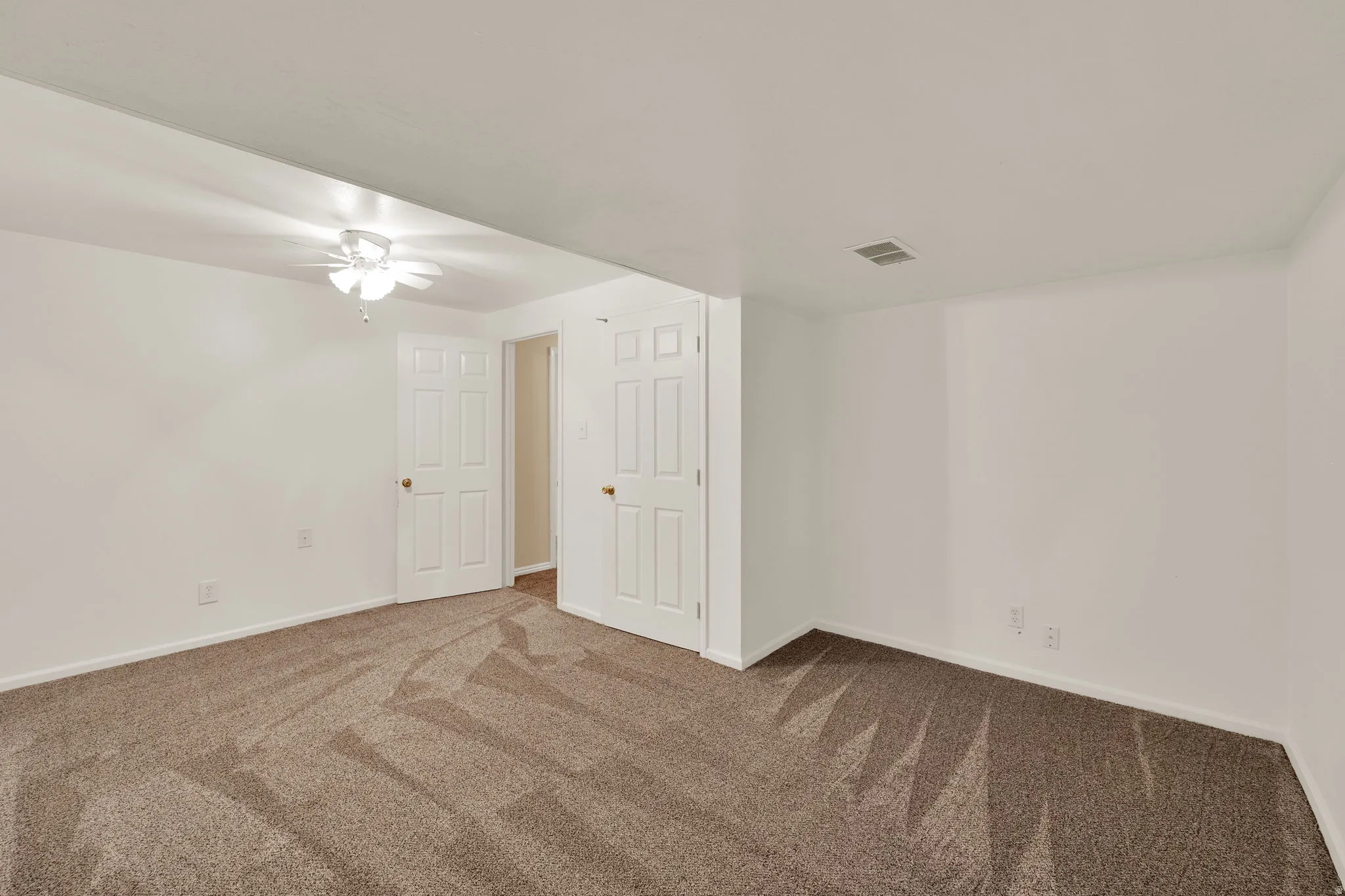 Carpeted room featuring white walls, a ceiling fan with light fixture, and two paneled doors with brass hardware