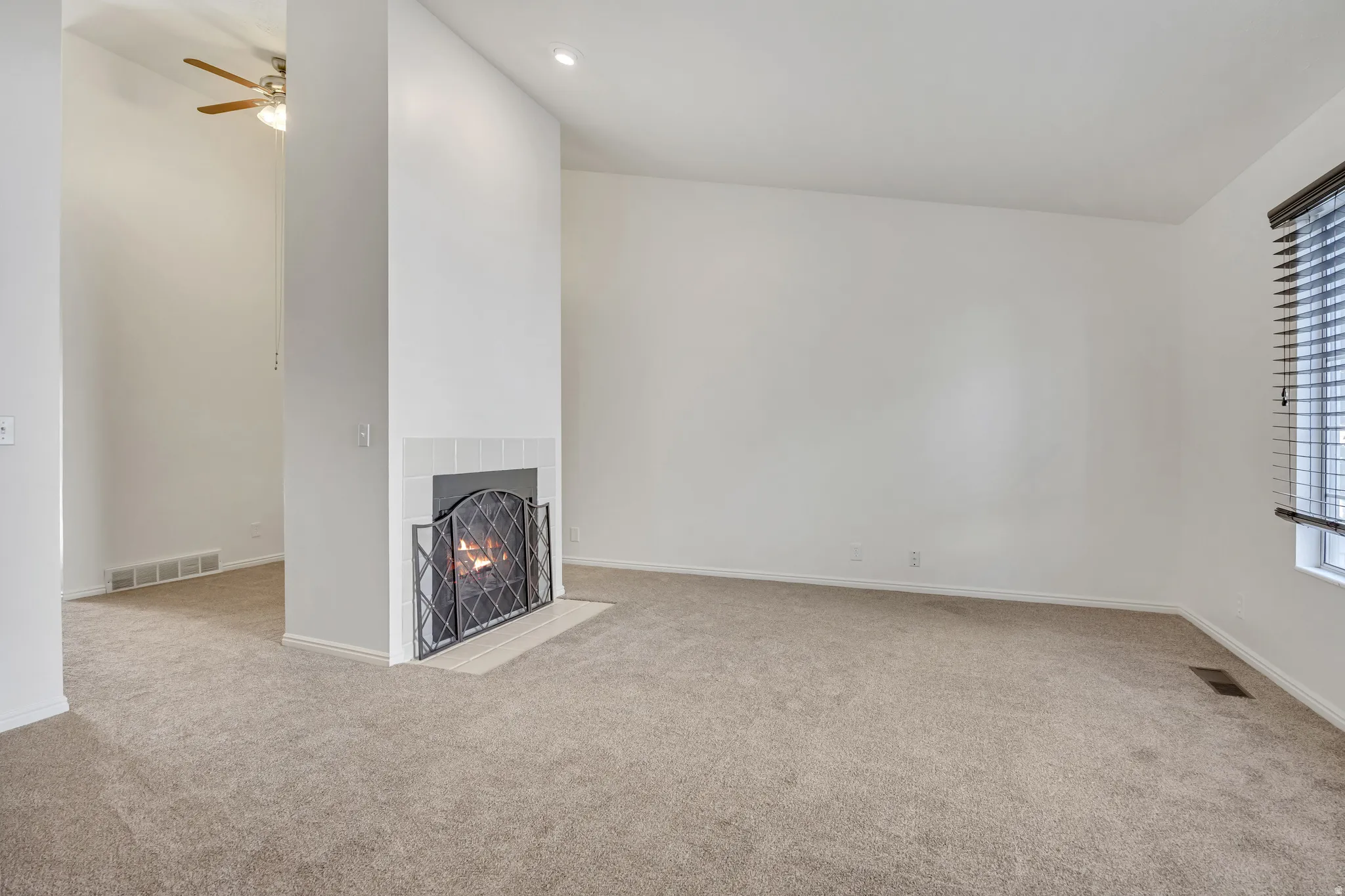 Spacious room featuring a fireplace with a white tile surround and hearth, neutral carpeting, a window with blinds, and a recessed ceiling light
