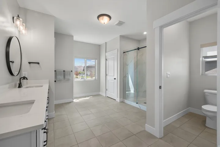Spacious bathroom featuring a double vanity with stone-finish countertops, a glass-enclosed shower, neutral tile flooring, and a separate water closet