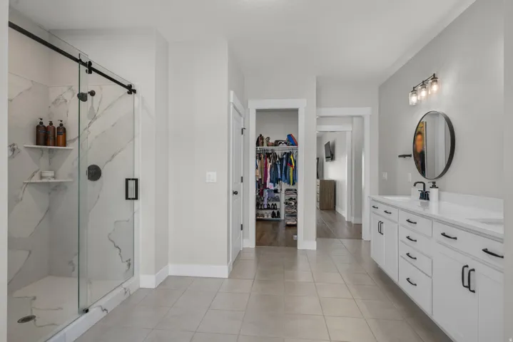 Spacious bathroom featuring a frameless glass shower with marble-look tile, a double vanity with white cabinetry and dark hardware, and large format floor tile