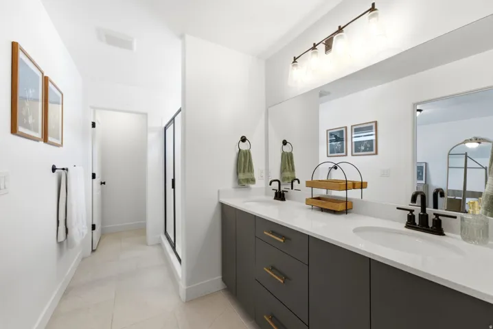 Dual vanity with white countertops and matte black fixtures, dark gray cabinetry with gold-tone hardware, full-width mirror, and tiled flooring