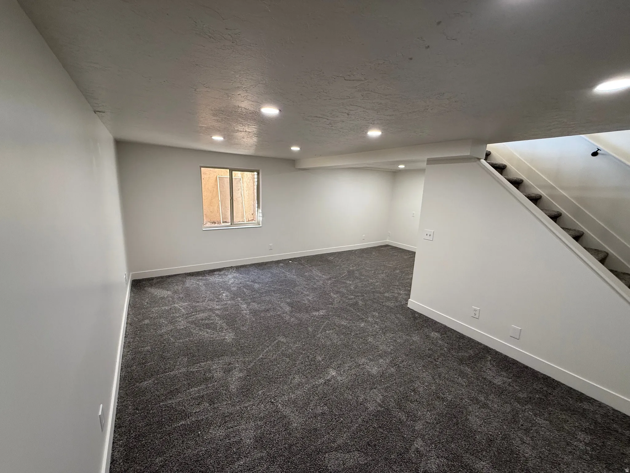Spacious interior room featuring plush gray carpeting, recessed ceiling lighting, and a window providing natural light
