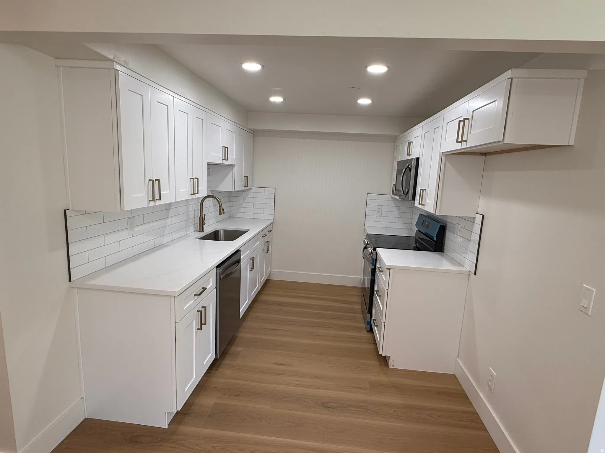 Galley-style kitchen featuring white shaker cabinetry, quartz countertops, a subway tile backsplash, stainless steel appliances, and wood-finish flooring