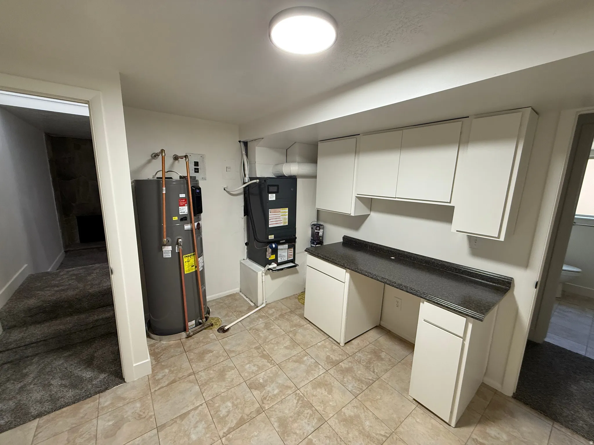Utility space featuring light-colored tile flooring, white cabinetry, dark countertop, and exposed water heater