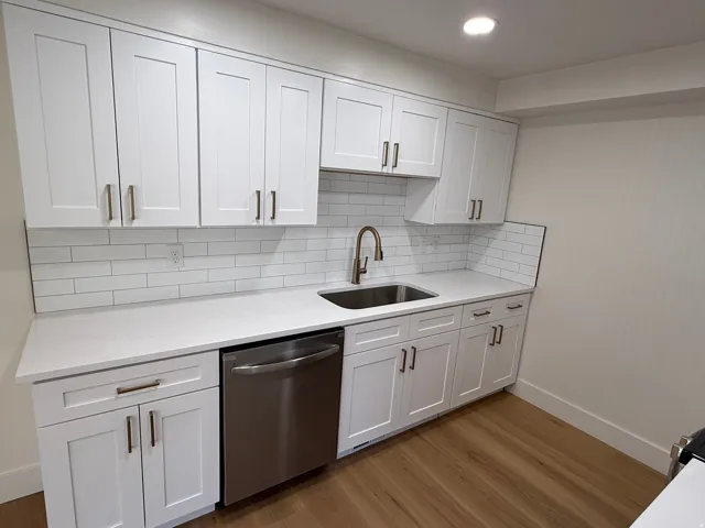 Modern kitchen featuring white shaker cabinetry, white subway tile backsplash, stainless steel sink with a gooseneck faucet, light-toned countertops, and wood-finish flooring