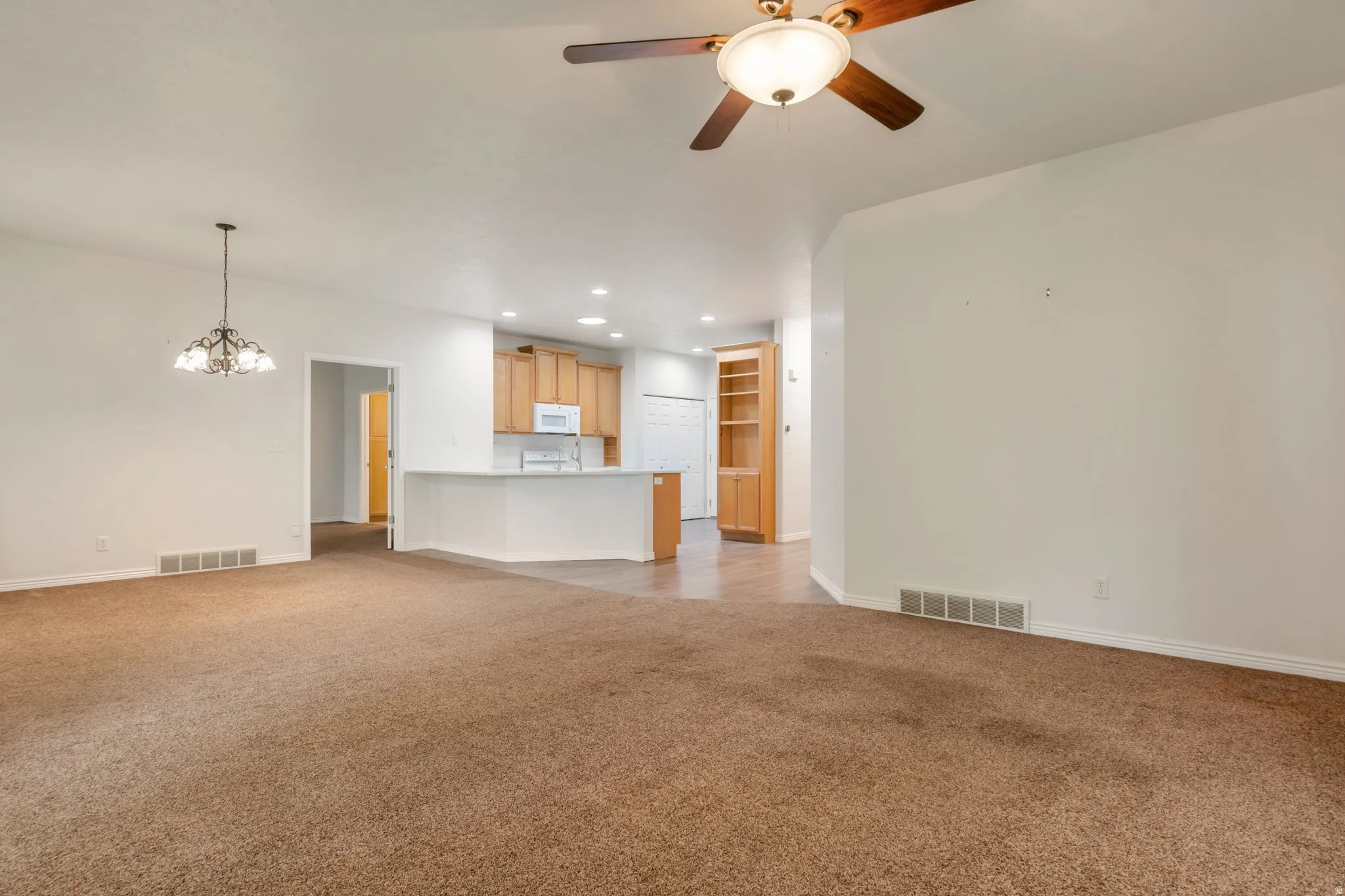 Open-concept living space featuring carpeted flooring, a ceiling fan with integrated lighting, a chandelier, a kitchen with light wood cabinetry, and a peninsula counter