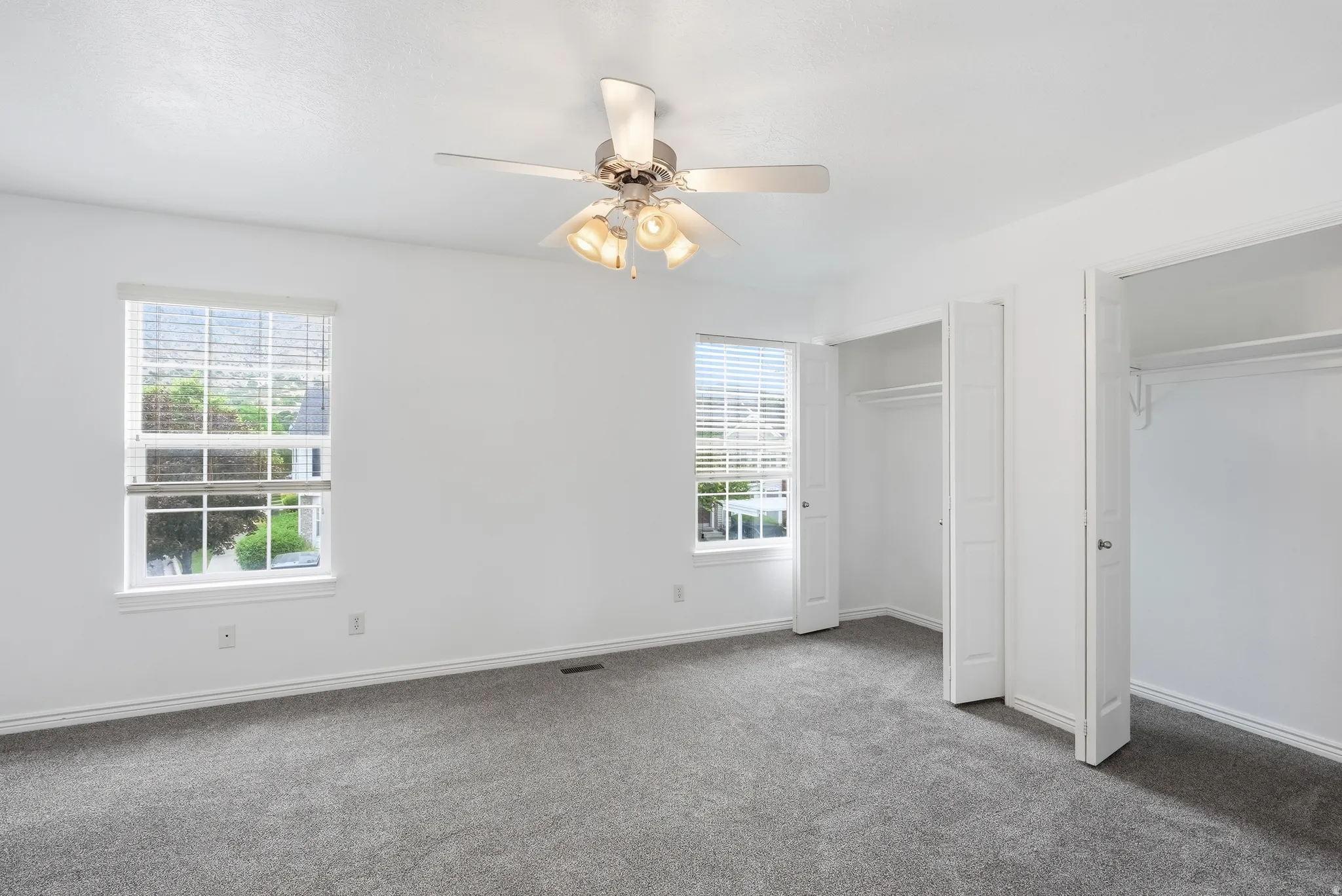 Light-filled room featuring plush gray carpeting, white walls, two window openings with blinds, a ceiling fan with integrated lighting, and two closet openings