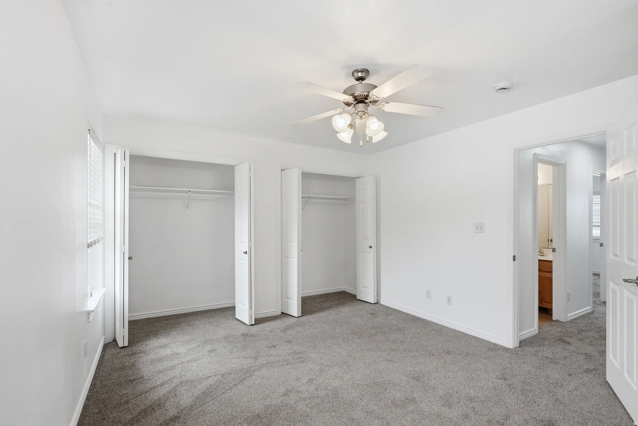 Spacious room featuring neutral carpeting, two double-door closets with shelving, a ceiling fan with integrated lighting, and a window with blinds