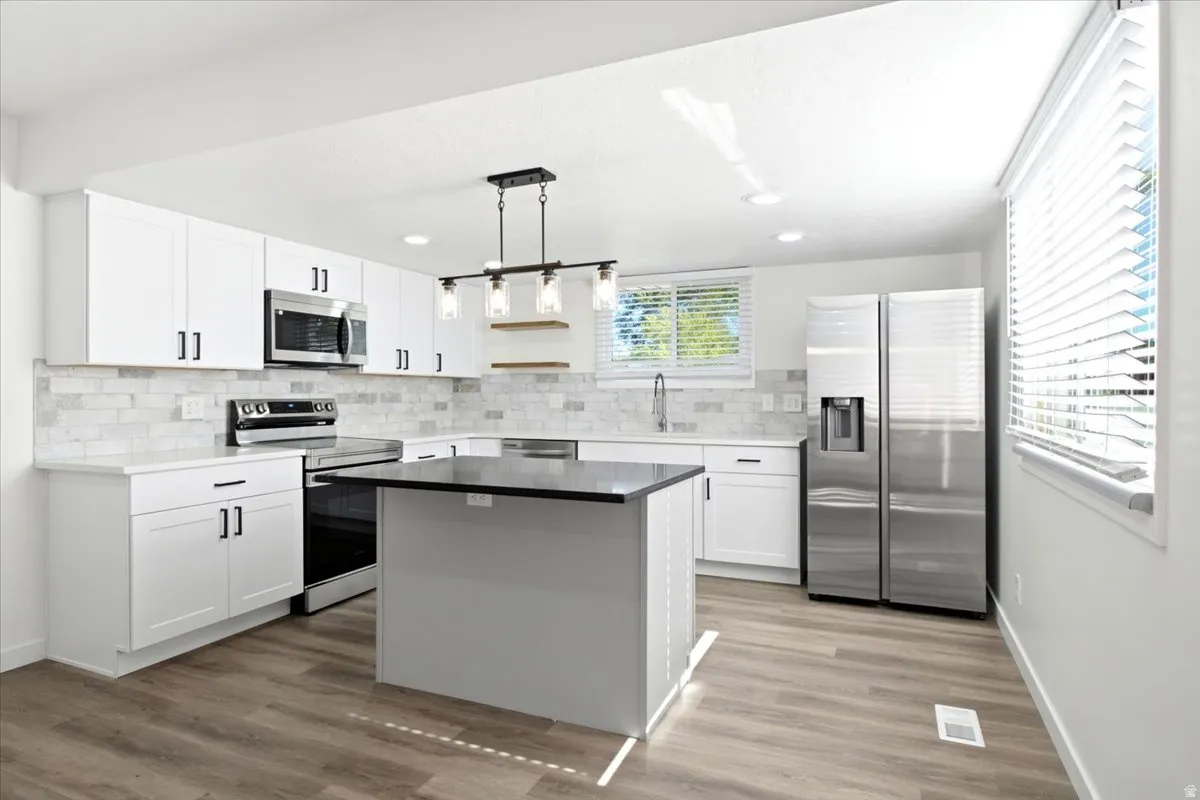 Kitchen featuring white shaker cabinetry, stainless steel appliances, a center island with a dark countertop, wood-finish flooring, and a subway tile backsplash