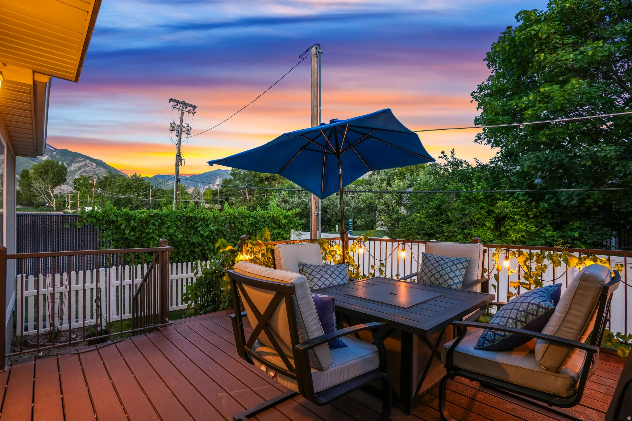 Expansive wood-finish deck featuring a built-in fire table, white picket fencing, and string lighting