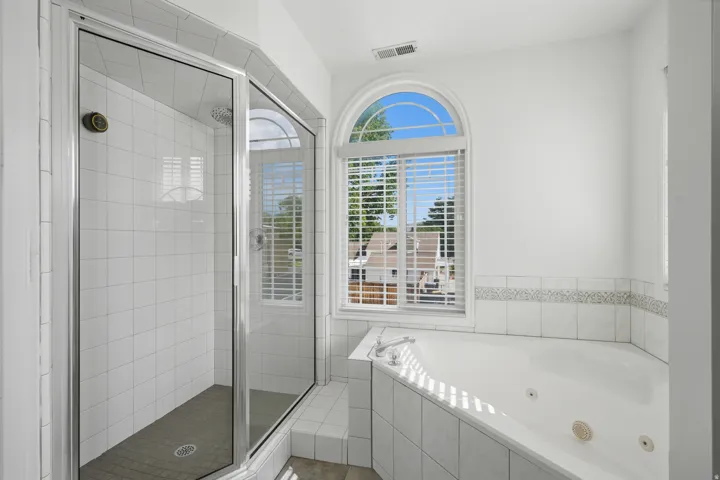 Spacious bathroom featuring a glass-enclosed shower with white subway tile, a built-in jetted tub with a tiled surround, and an arched window with blinds