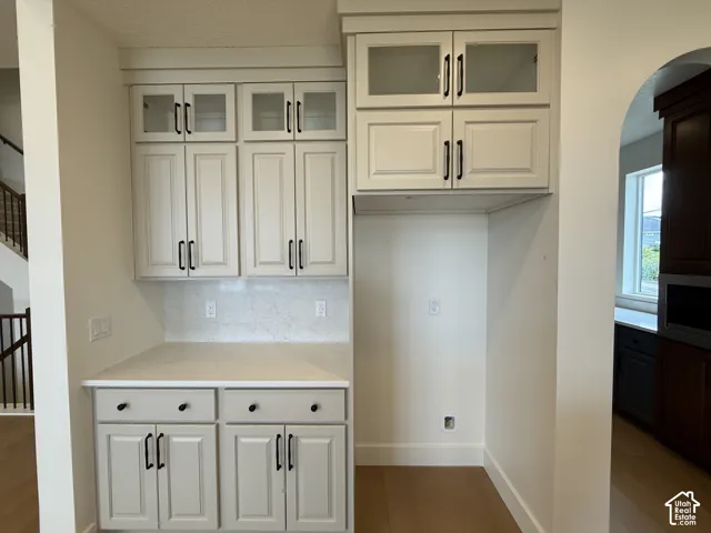 Off-white cabinetry featuring glass-front upper displays, matte black hardware, light-toned stone-finish countertops, and an arched entryway