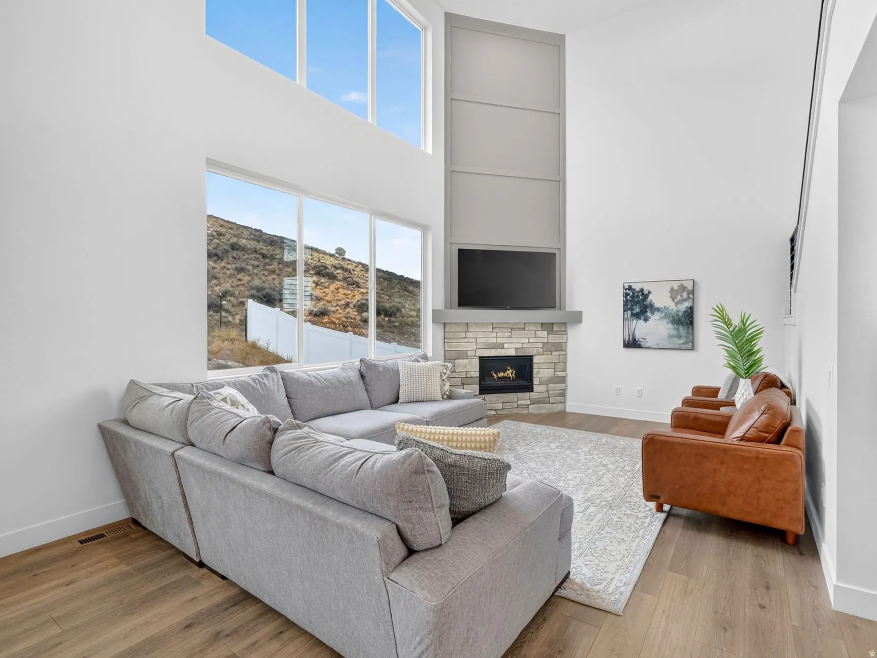 Two-story living area featuring tall ceilings, expansive windows, a stacked stone fireplace, wood-finish flooring, and a modern black metal railing