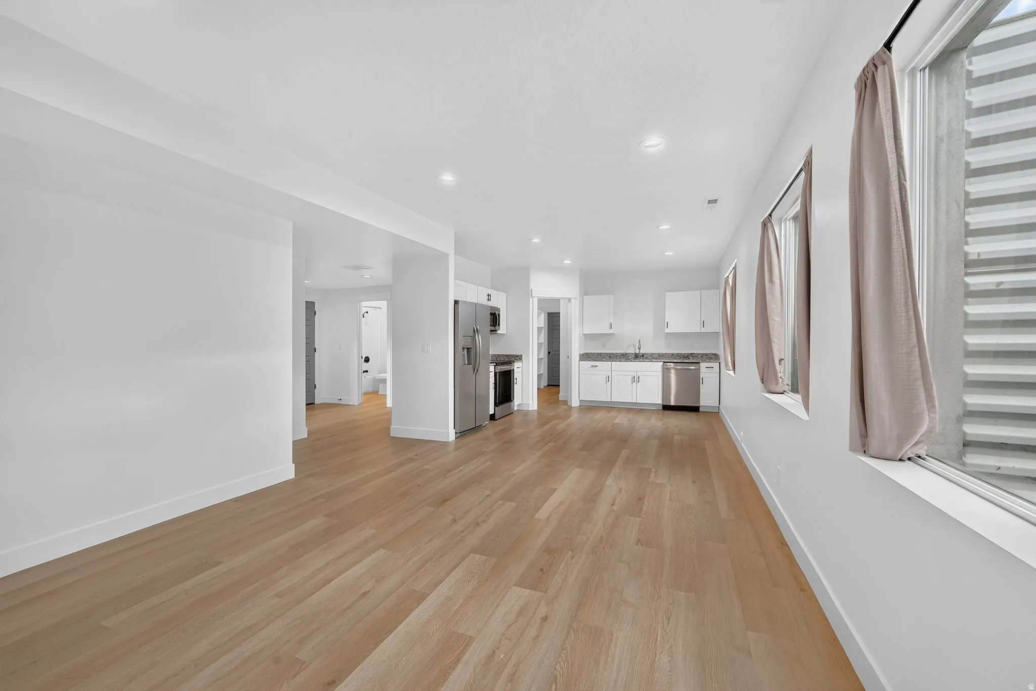 Open-concept living area featuring light wood-finish flooring, recessed lighting, and a kitchen with white cabinetry, stainless steel appliances, and stone countertops