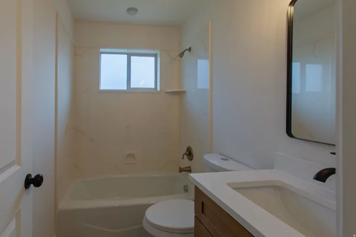 Bathroom featuring a white bathtub with a window above, white shower surround, a vanity with a white countertop and rectangular sink, and a dark-framed mirror