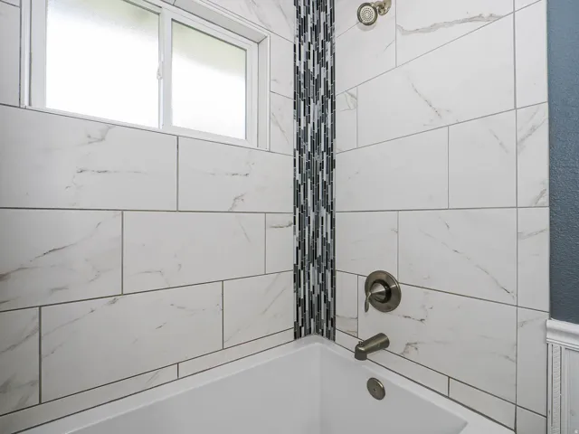 Built-in bathtub featuring a brushed nickel faucet and showerhead, white rectangular wall tiles with gray veining, a vertical mosaic tile accent, and a frosted window