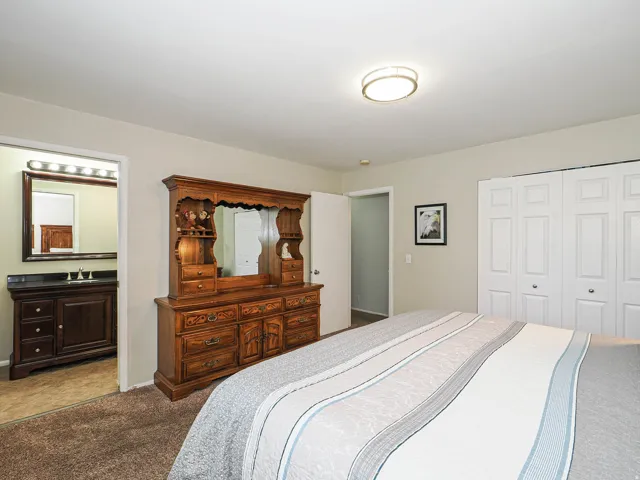Carpeted room with a ceiling-mounted light fixture, bi-fold closet doors, and an open doorway revealing a bathroom with a vanity, sink, and mirror