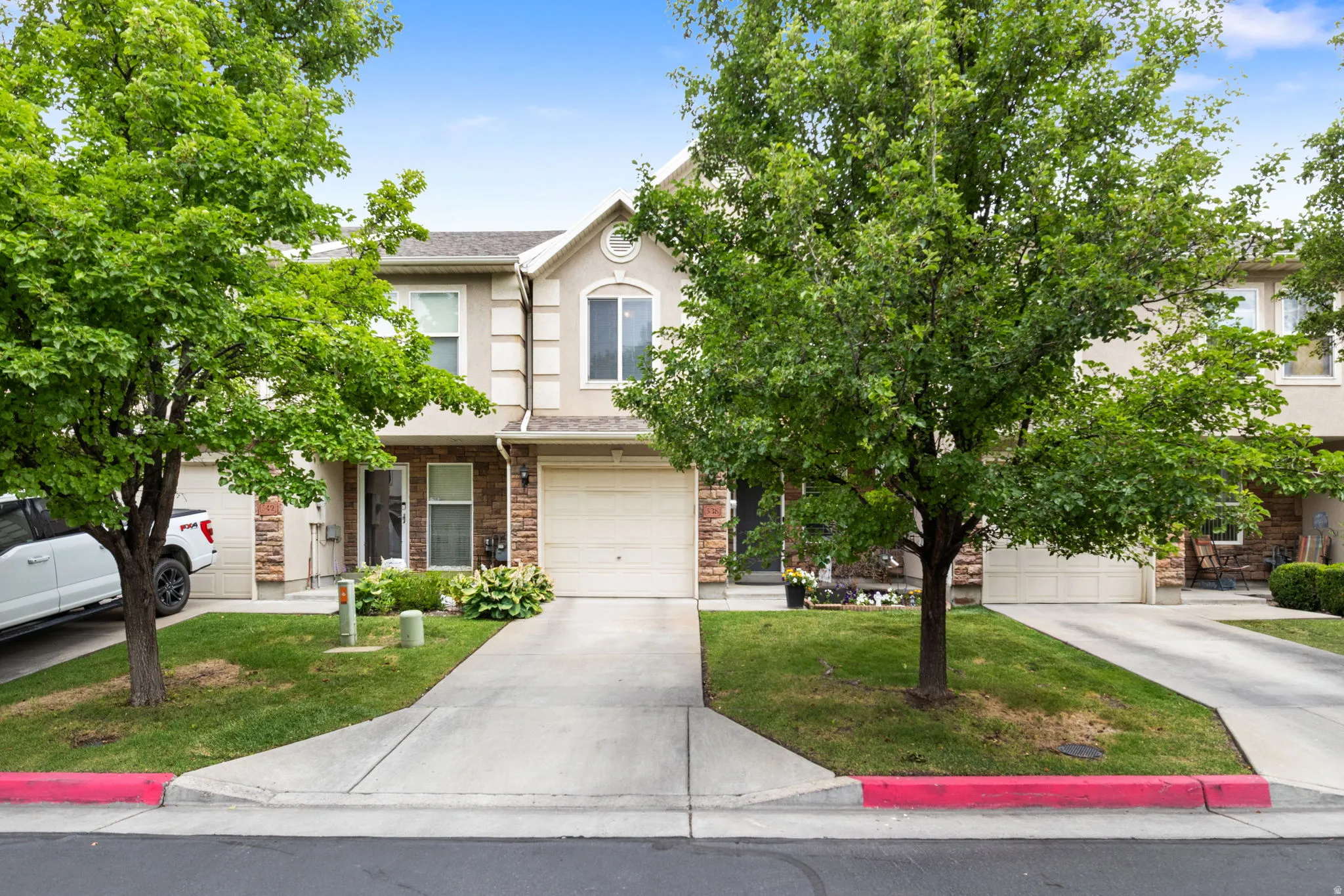 Townhome exterior featuring a stucco and stone facade, an attached garage, a concrete driveway, and mature trees
