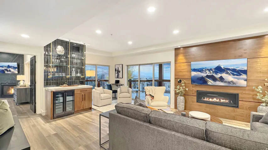 Spacious living area featuring wood-finish flooring, a linear electric fireplace set into a wood-paneled wall, a built-in wet bar with beverage coolers, and extensive glass doors