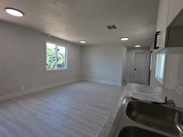 Open-concept living area featuring light wood-finish flooring, recessed ceiling lighting, and a kitchen area with a double basin stainless steel sink and light-toned countertop