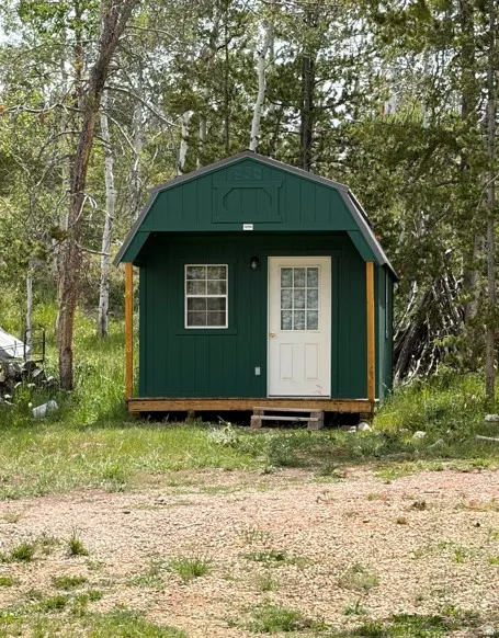 Green gambrel-roofed structure with a white entry door and a single grid-paned window