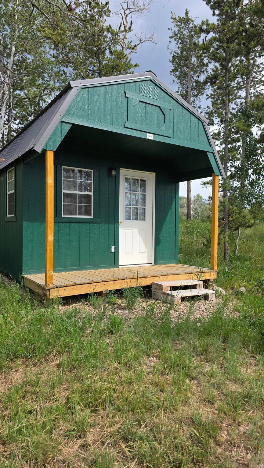Green-sided structure featuring a gambrel roof and a covered front porch
