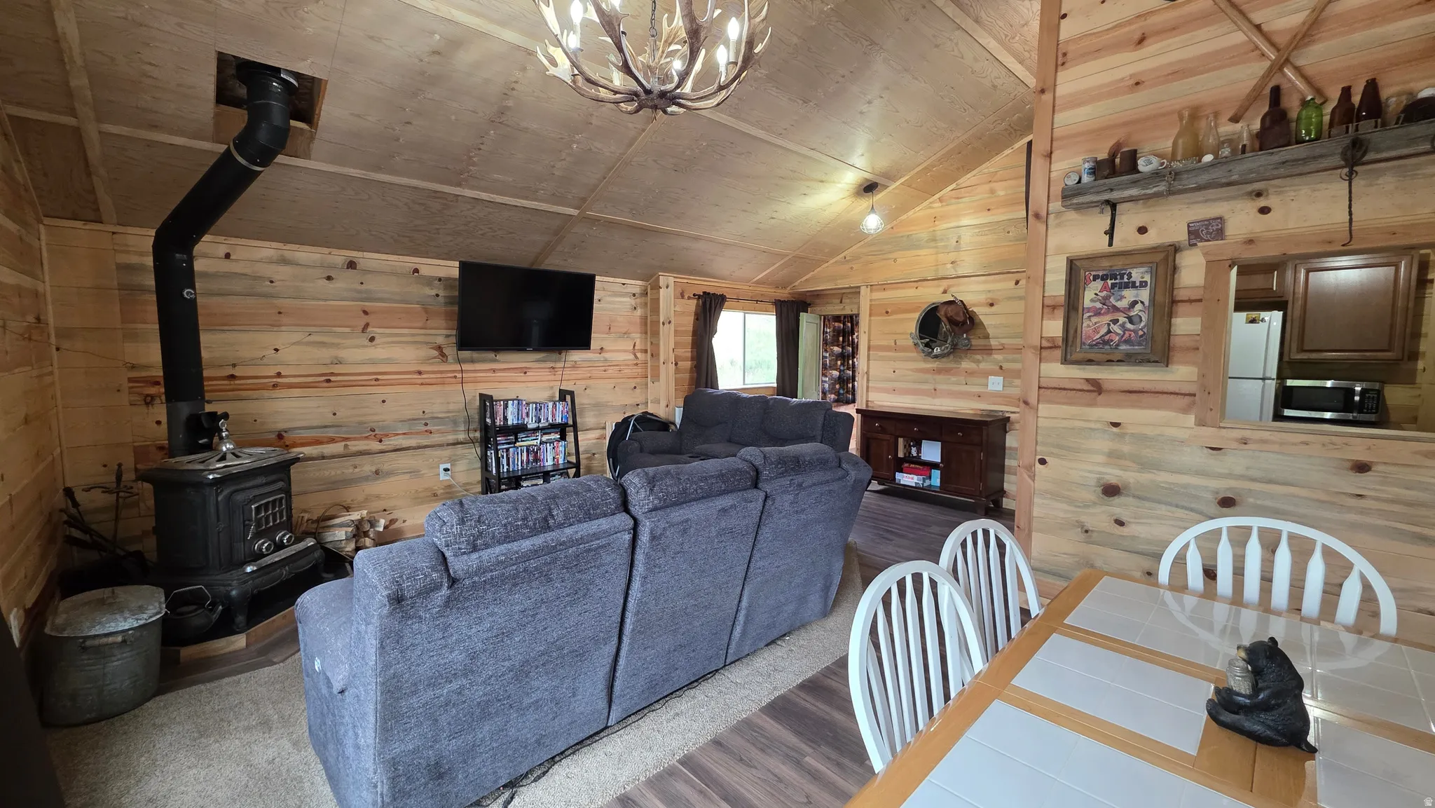 Open-concept living space featuring wood-paneled walls, vaulted ceilings, and a prominent wood-burning stove with an exposed flue