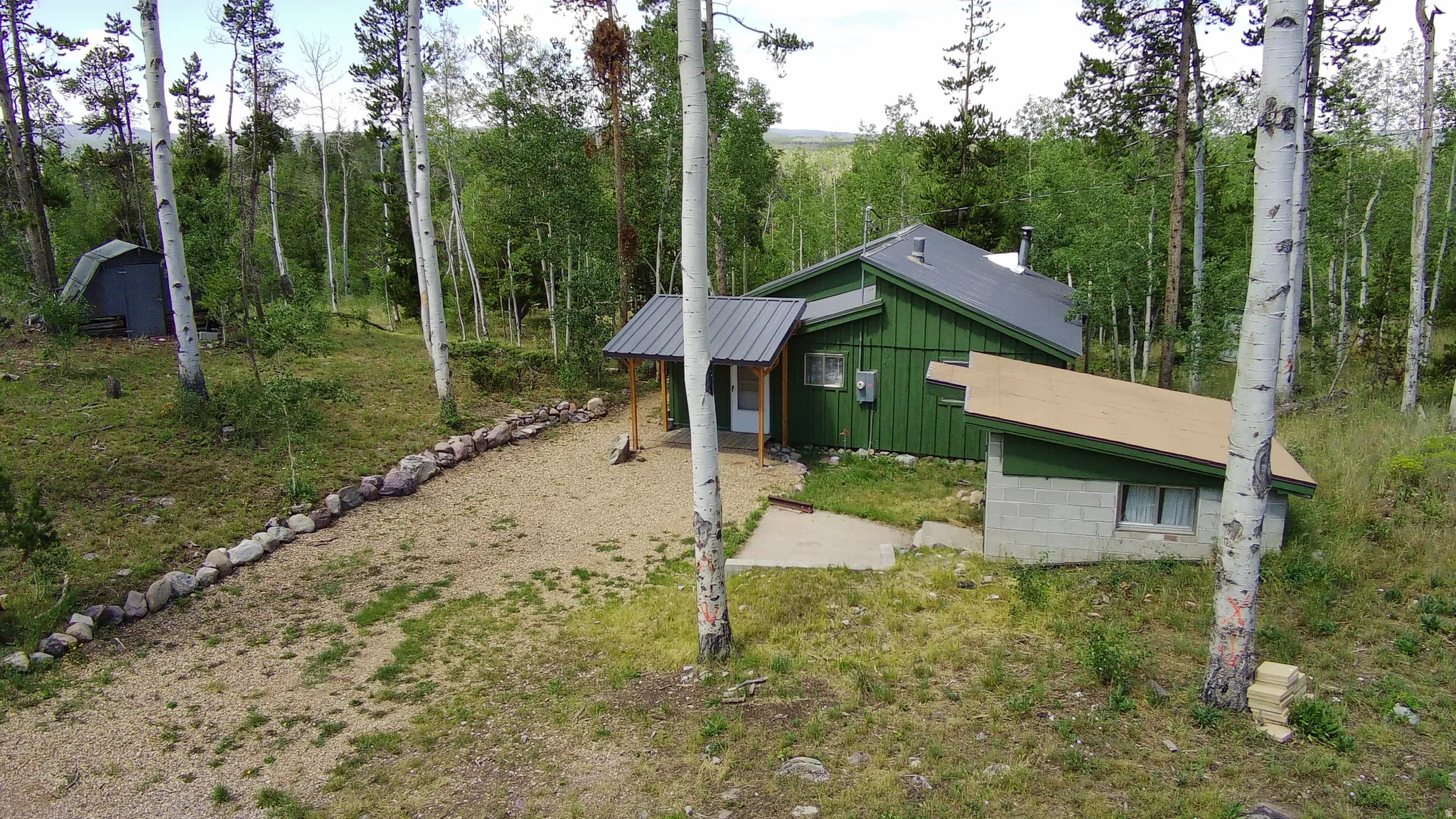 Wooded property featuring a green siding structure with a dark metal roof, a smaller auxiliary building, and a gravel driveway