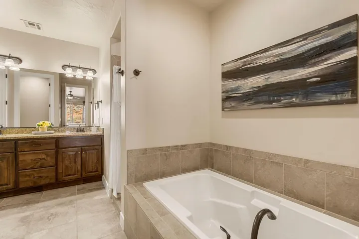 Soaking tub with tile surround, dual vanity with granite-style countertop, dark wood-finish cabinetry, and large mirror