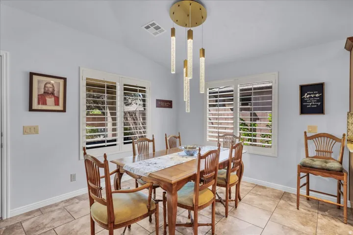 Dining area featuring a modern gold-finish chandelier with cylindrical lights, two windows with white plantation shutters, tile flooring, and light-colored walls