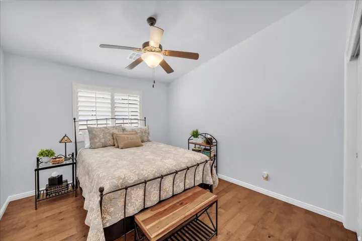 Bedroom featuring wood-finish flooring, a ceiling fan with light fixture, and window with white plantation shutters