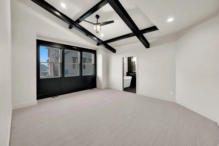 Spacious room featuring a vaulted ceiling with exposed black beams, light-toned carpeting, and a large multi-pane window with black framing