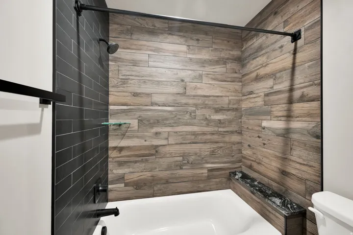 Bathtub shower featuring wood-look tile surround, dark subway tile accent, a built-in bench with a dark countertop, and a rainfall showerhead