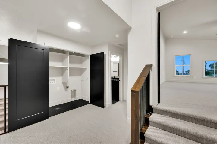 Upper-level landing featuring a laundry area with black hexagon floor tiles and built-in shelving, a wood and metal stair railing, and a carpeted room with multiple windows