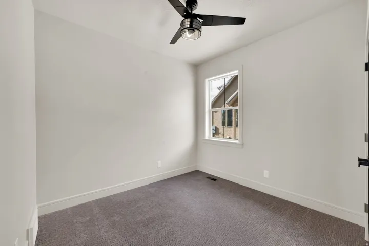 Interior room featuring neutral wall paint, gray patterned carpeting, a window with white trim, a black ceiling fan with an integrated light fixture, and white baseboards