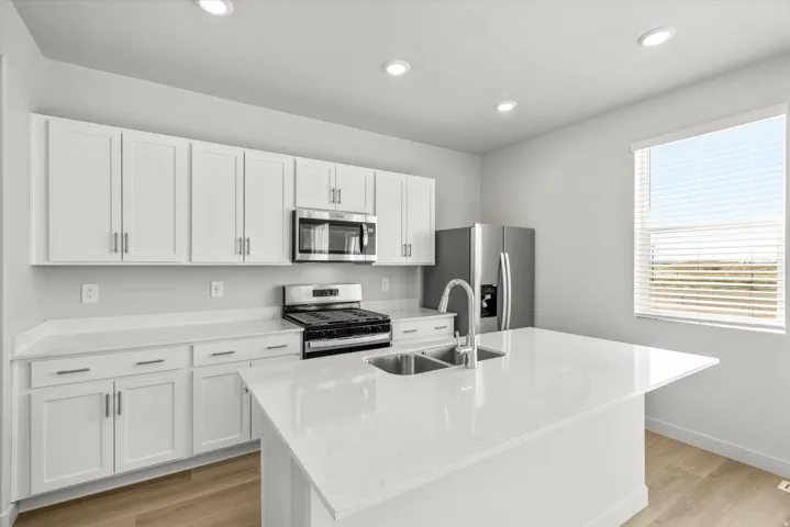 Modern kitchen featuring white shaker-style cabinetry, stainless steel appliances including a gas cooktop and microwave, white quartz countertops, an oversized island with waterfall edge, chrome faucet with pull-down sprayer, recessed ceiling lighting, an