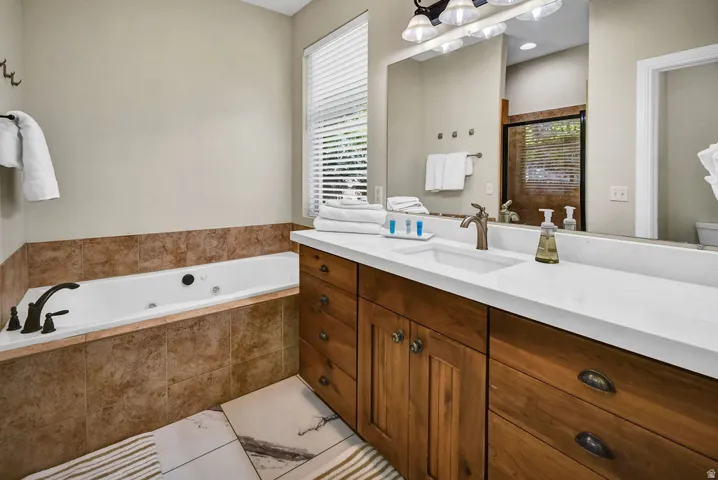 Spacious bathroom featuring a built-in jetted tub with tile surround, a double vanity with ample storage, a large wall-mounted mirror, and a window with privacy blinds