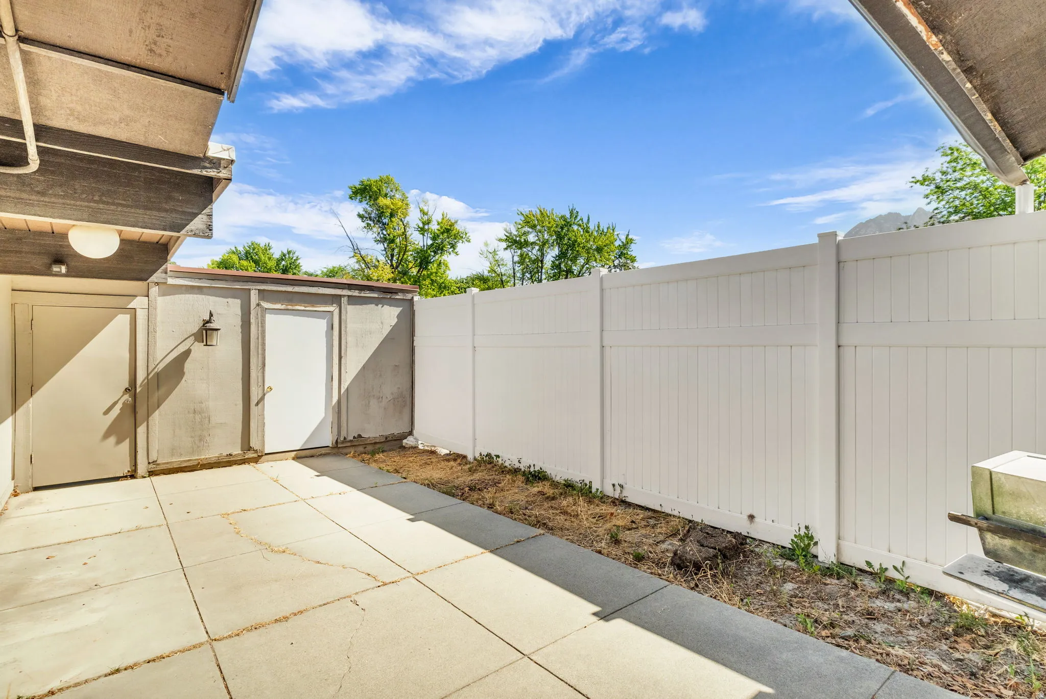 Concrete patio area featuring a built-in storage shed with an exterior light fixture, and a durable white privacy fence
