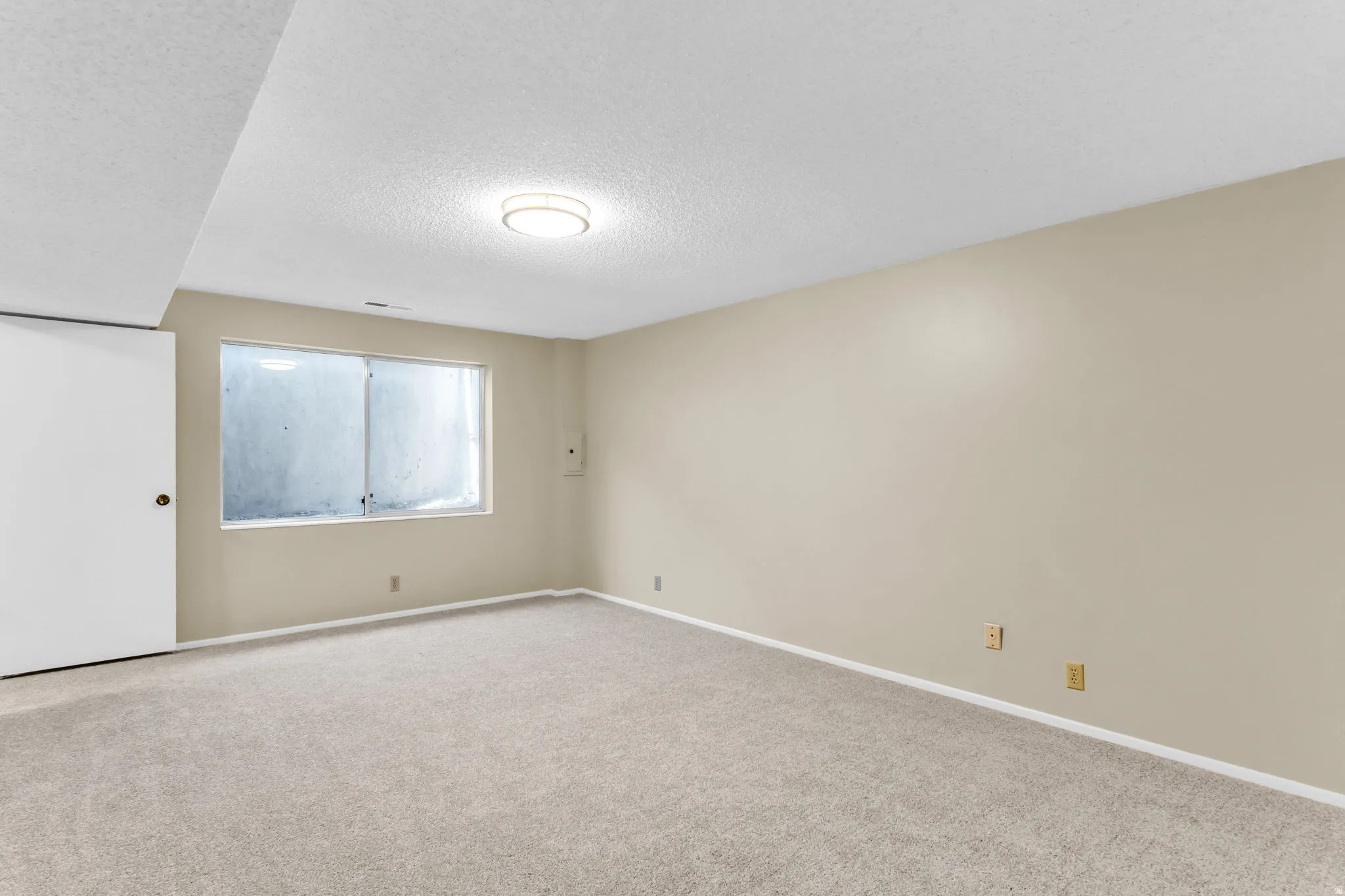 Spacious room featuring neutral carpet flooring, light beige wall paint, a large window, a flush-mount ceiling light fixture, and white baseboards