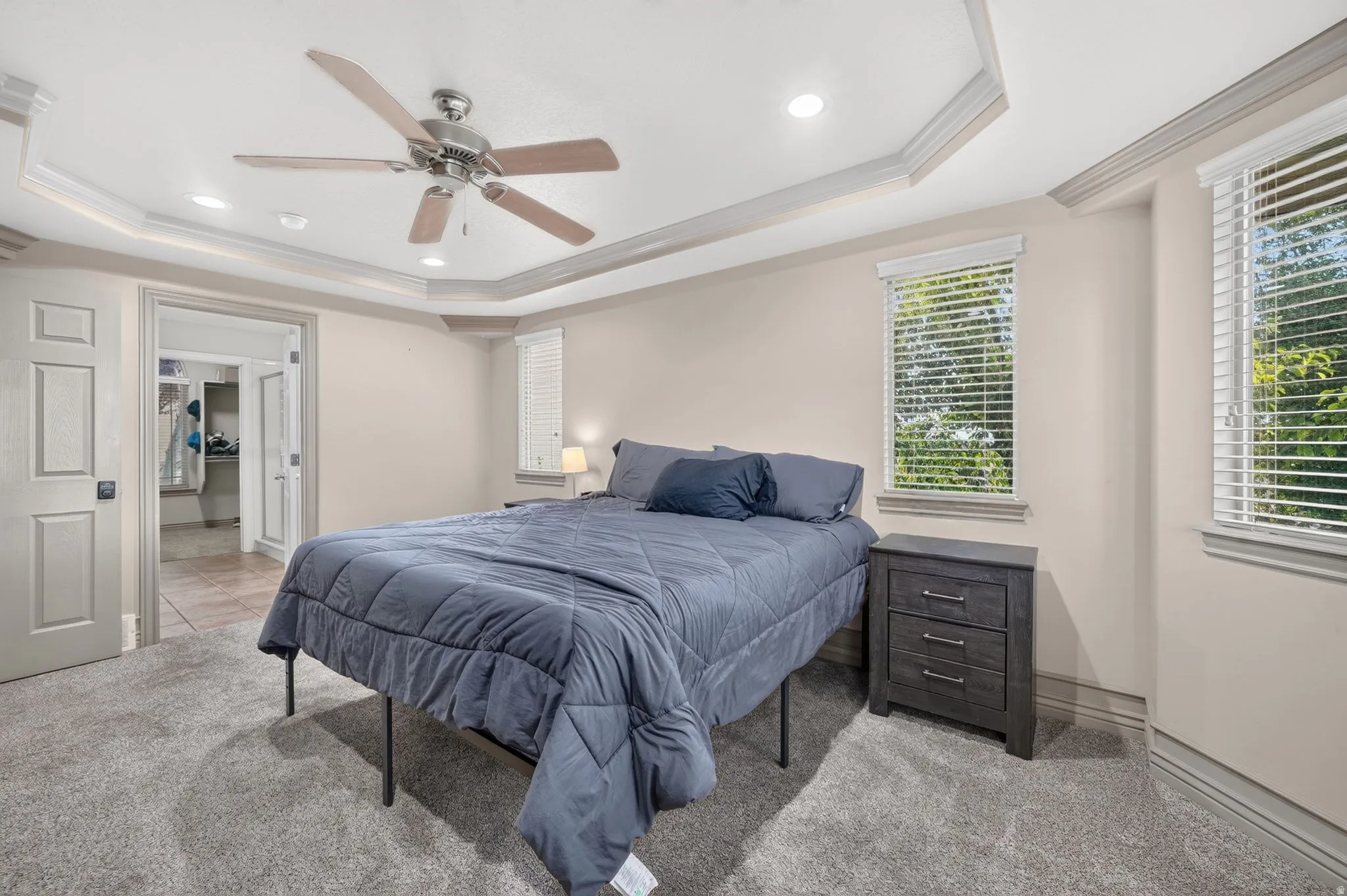 Spacious bedroom featuring plush gray carpeting, two windows with white blinds, a tray ceiling with a ceiling fan, and recessed lighting