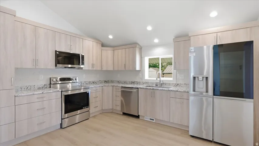Modern kitchen featuring light wood-finish cabinetry, stainless steel appliances, granite countertops, and a white tile backsplash